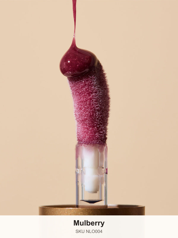 Glossy deep berry INIKA lip oil pouring onto a fuzzy sponge applicator above a gold cap, forming a rounded droplet over the tip. The caption identifies the shade as Mulberry.