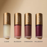 Four frosted INIKA lip oil bottles with gold caps lined up on a beige ledge, labelled left to right: Clear, peach-pink Blossom, reddish-brown Cinnamon and deep berry Mulberry.