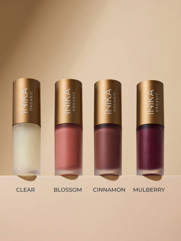 Four frosted INIKA lip oil bottles with gold caps lined up on a beige ledge, labelled left to right: Clear, peach-pink Blossom, reddish-brown Cinnamon and deep berry Mulberry.