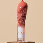 Extreme close-up of glossy coral-pink INIKA lip oil pouring onto a fuzzy sponge applicator above a gold cap, forming a thick droplet over the tip.