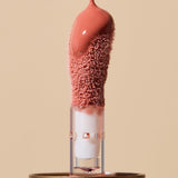 Extreme close-up of glossy coral-pink INIKA lip oil pouring onto a fuzzy sponge applicator above a gold cap, forming a thick droplet over the tip.