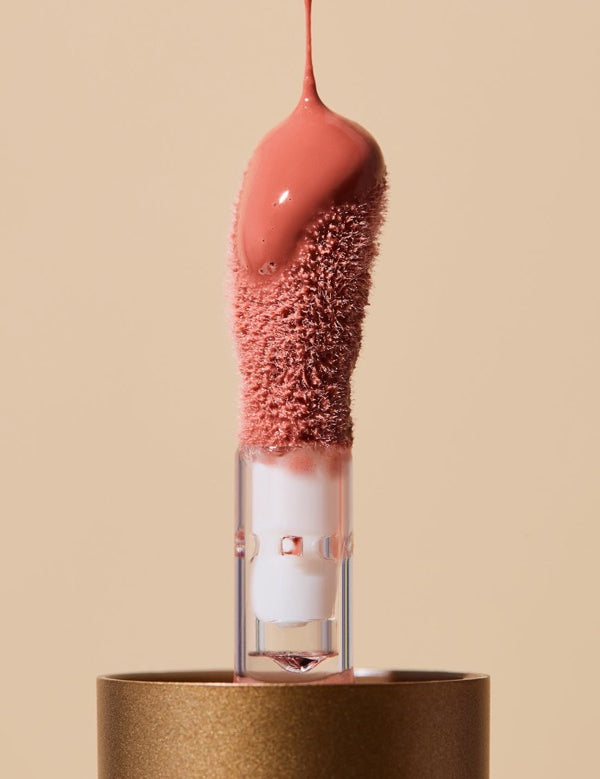 Extreme close-up of glossy coral-pink INIKA lip oil pouring onto a fuzzy sponge applicator above a gold cap, forming a thick droplet over the tip.