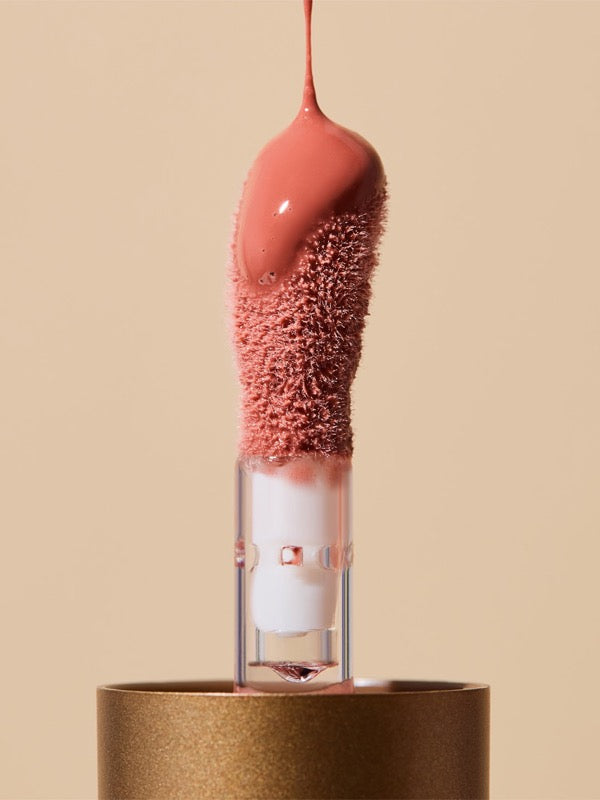 Extreme close-up of glossy coral-pink INIKA lip oil pouring onto a fuzzy sponge applicator above a gold cap, forming a thick droplet over the tip.