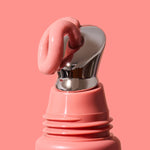 Thick glossy pink INIKA lip serum curling over the slanted silver applicator on a coral-pink tube against a matching background. The caption identifies the shade as Blush.