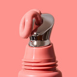 Thick glossy pink INIKA lip serum curling over the slanted silver applicator on a coral-pink tube against a matching background. The caption identifies the shade as Blush.