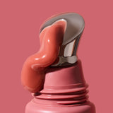 Thick glossy rose-pink INIKA lip serum curling down the slanted silver applicator on a dusty-pink tube against a matching background. The caption identifies the shade as Peony.