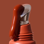 Thick glossy red-brown INIKA lip serum flowing down the slanted silver applicator on a terracotta tube against a brown background. The caption identifies the shade as Rosewood.