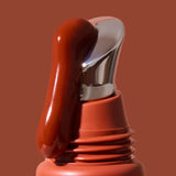 Thick glossy red-brown INIKA lip serum flowing down the slanted silver applicator on a terracotta tube against a brown background. The caption identifies the shade as Rosewood.
