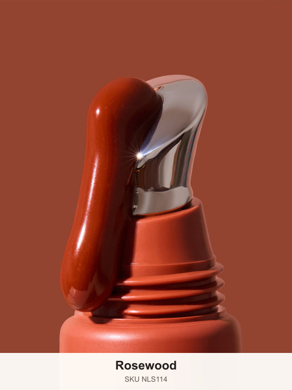 Thick glossy red-brown INIKA lip serum flowing down the slanted silver applicator on a terracotta tube against a brown background. The caption identifies the shade as Rosewood.