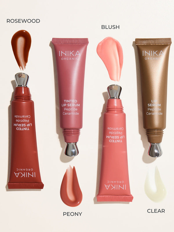 Four uncapped INIKA lip serum tubes arranged beside glossy swatches on a pale background, labelled Rosewood with red-brown serum, Peony with rose-pink serum, Blush with light pink serum and Clear with translucent serum.