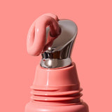 Extreme close-up of thick glossy pink INIKA lip serum curling over the slanted silver applicator on a coral-pink tube against a matching pink background.