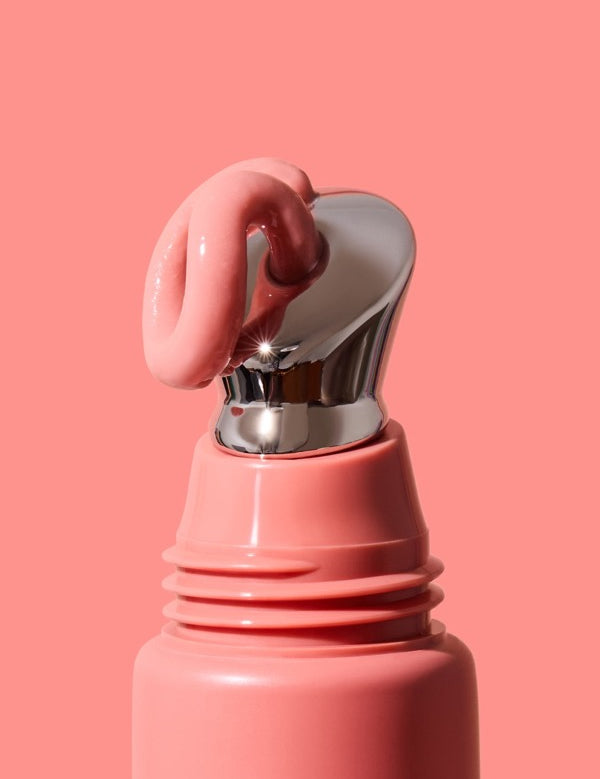 Extreme close-up of thick glossy pink INIKA lip serum curling over the slanted silver applicator on a coral-pink tube against a matching pink background.