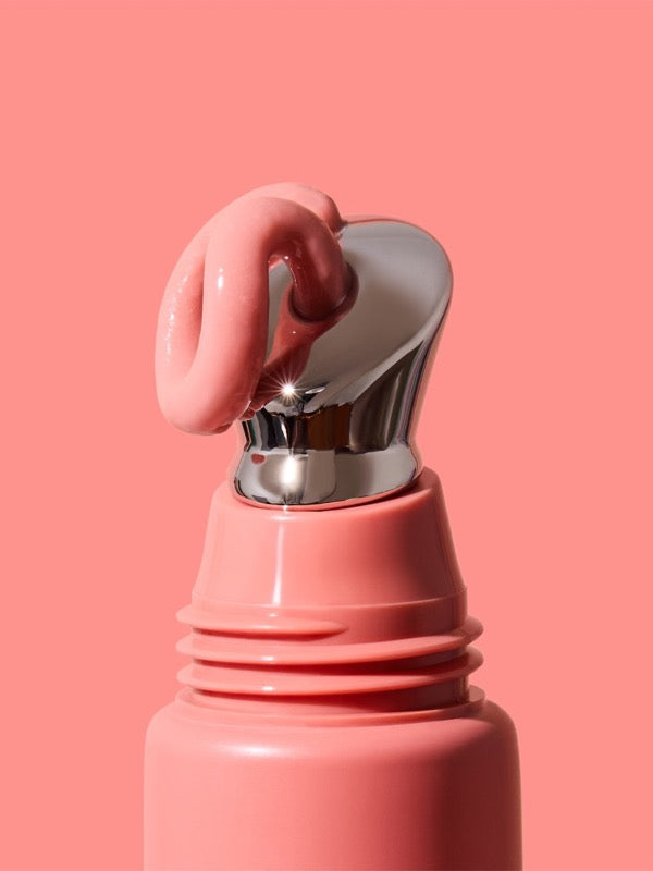 Extreme close-up of thick glossy pink INIKA lip serum curling over the slanted silver applicator on a coral-pink tube against a matching pink background.
