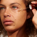Close-up of a person with light brown curls holding a glass dropper beside their cheek, with a golden bead of INIKA Phyto-Active Face Oil at its tip.