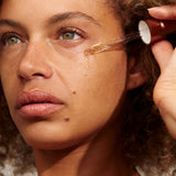 Close-up of a person with light brown curls holding a glass dropper beside their cheek, with a golden bead of INIKA Phyto-Active Face Oil at its tip.