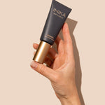 Hand holding a black and gold tube of INIKA Organic Pure Perfection Primer against a warm beige background with a sharp natural shadow.