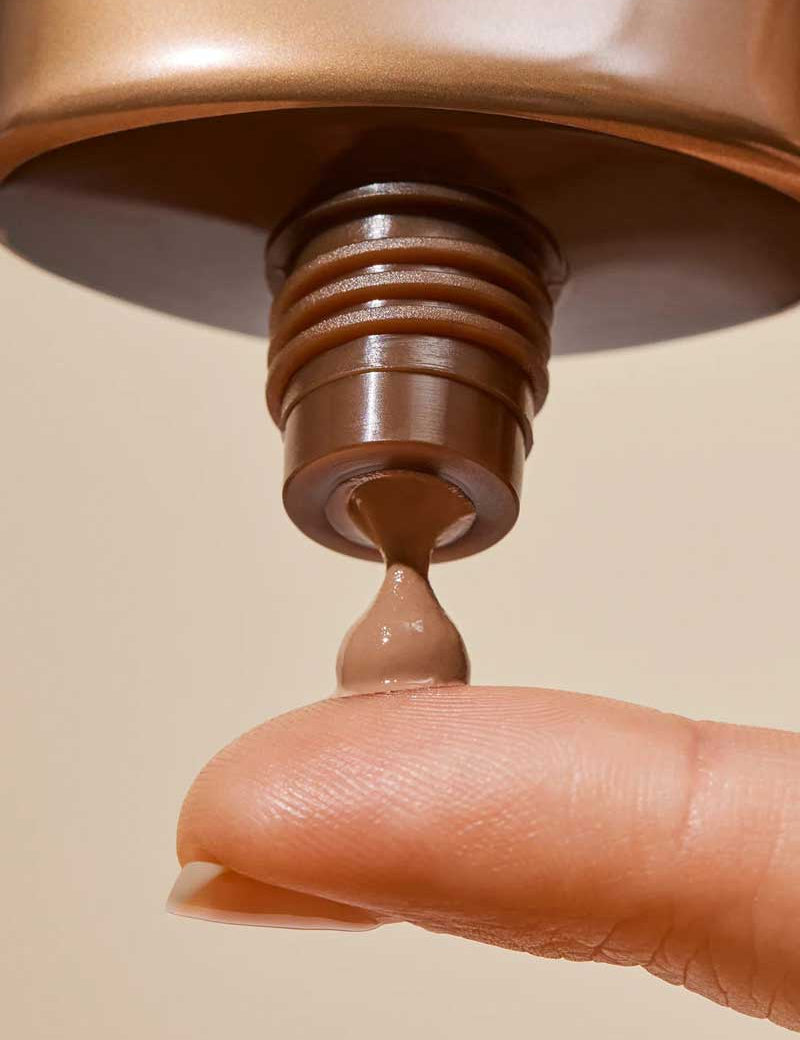 A close-up shows a finger catching a small amount of INIKA Organic Tinted Natural Sunscreen SPF50+ 50ml being dispensed from its tube against a neutral background.
