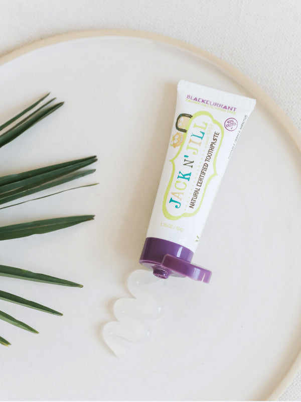 Open Jack N' Jill Blackcurrant toothpaste tube on a round white tray beside a palm frond and a wavy line of translucent paste.