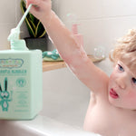 Child sitting in a white bathtub and raising a long green bubble wand above a mint-green Jack N' Jill Blissful Bubbles bottle on the bath ledge.