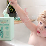 Child sitting in a white bathtub and raising a long green bubble wand above a mint-green Jack N' Jill Blissful Bubbles bottle on the bath ledge.