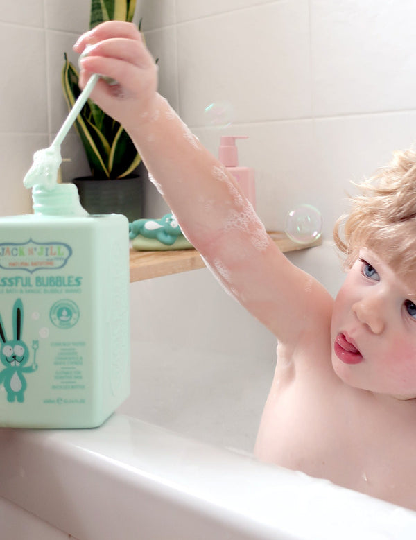 Child sitting in a white bathtub and raising a long green bubble wand above a mint-green Jack N' Jill Blissful Bubbles bottle on the bath ledge.