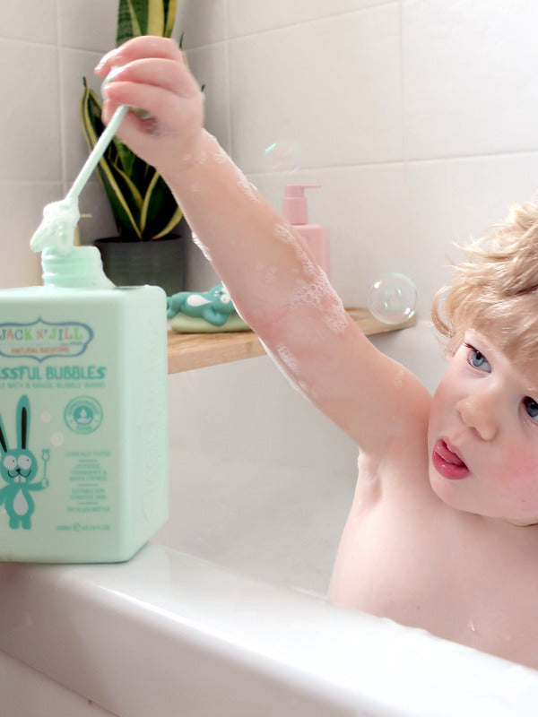 Child sitting in a white bathtub and raising a long green bubble wand above a mint-green Jack N' Jill Blissful Bubbles bottle on the bath ledge.