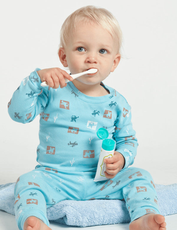 Young child in turquoise pyjamas brushing their teeth while holding an open Jack N' Jill Blueberry toothpaste tube.