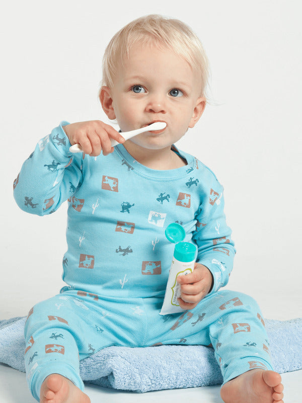 Young child in turquoise pyjamas brushing their teeth while holding an open Jack N' Jill Blueberry toothpaste tube.