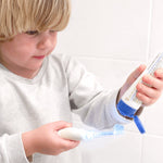 Child squeezing Jack N' Jill Bubblegum toothpaste from the open tube onto a toothbrush beside a white-tiled wall.