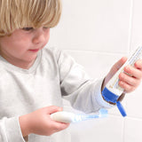 Child squeezing Jack N' Jill Bubblegum toothpaste from the open tube onto a toothbrush beside a white-tiled wall.