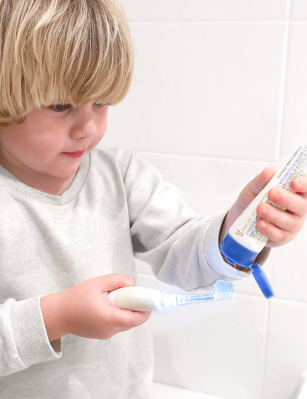 Child squeezing Jack N' Jill Bubblegum toothpaste from the open tube onto a toothbrush beside a white-tiled wall.