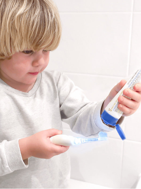 Child squeezing Jack N' Jill Bubblegum toothpaste from the open tube onto a toothbrush beside a white-tiled wall.