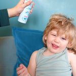 Jack N' Jill Detangler and Leave-in Conditioner being sprayed onto a smiling child's hair while the child sits on a blue sofa.