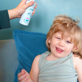 Jack N' Jill Detangler and Leave-in Conditioner being sprayed onto a smiling child's hair while the child sits on a blue sofa.