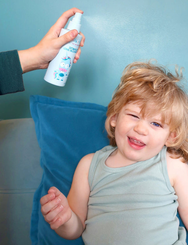 Jack N' Jill Detangler and Leave-in Conditioner being sprayed onto a smiling child's hair while the child sits on a blue sofa.