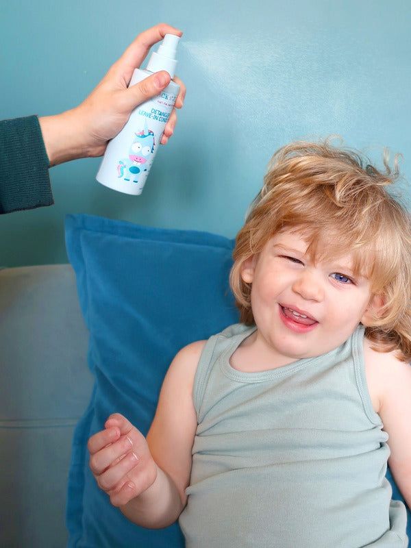 Jack N' Jill Detangler and Leave-in Conditioner being sprayed onto a smiling child's hair while the child sits on a blue sofa.