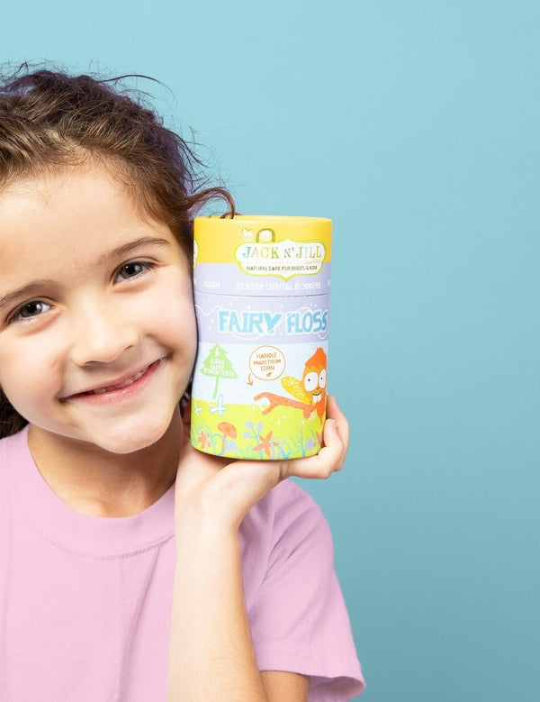 Smiling child holding a yellow box of Jack N' Jill Fairy Floss Picks beside her face against a soft blue background.