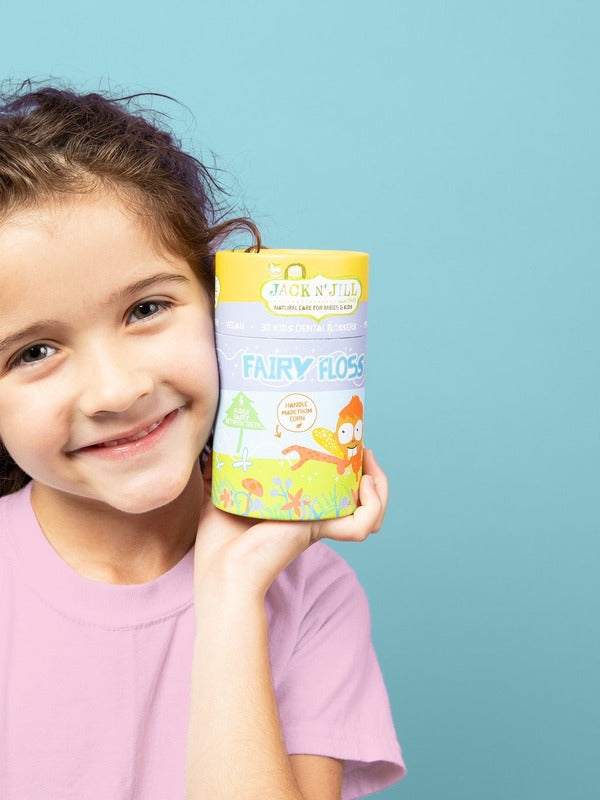 Smiling child holding a yellow box of Jack N' Jill Fairy Floss Picks beside her face against a soft blue background.