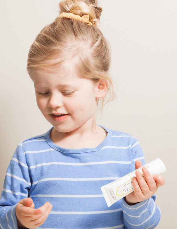 Child with blonde hair in a high bun holding a Jack N' Jill Flavour Free toothpaste tube while preparing to brush.