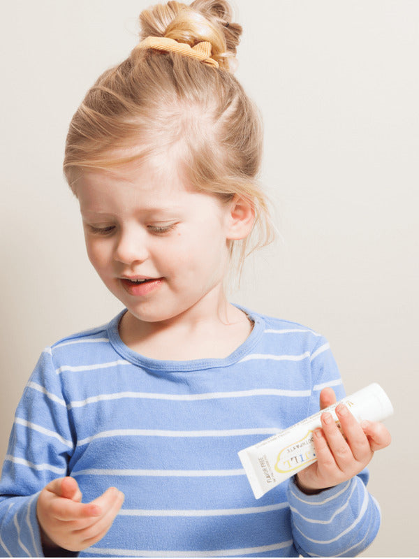 Child with blonde hair in a high bun holding a Jack N' Jill Flavour Free toothpaste tube while preparing to brush.