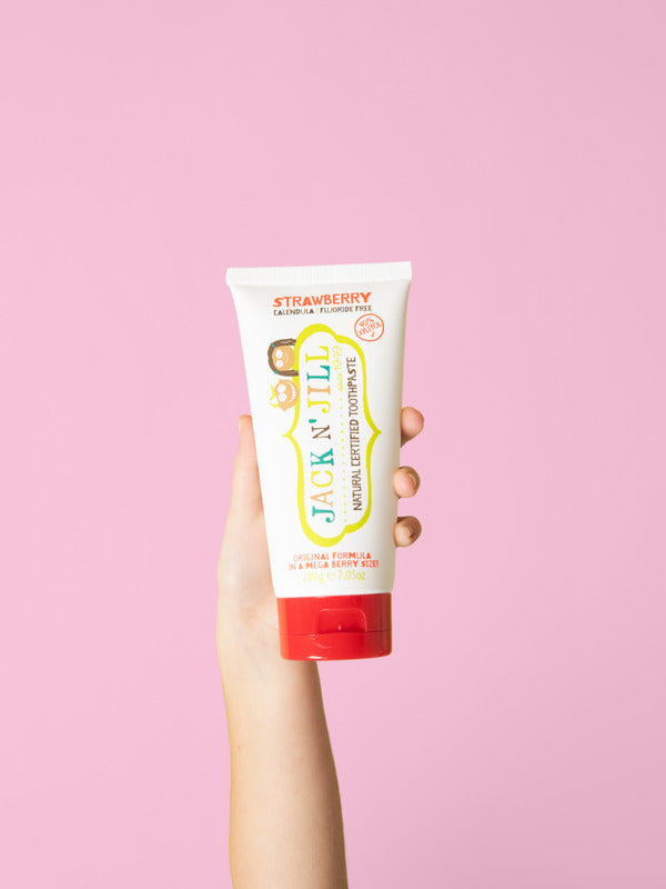 Hand holding the large Jack N' Jill Mega Berry Strawberry toothpaste tube upright against a bright pink background.
