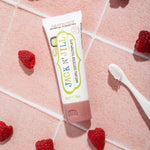 Jack N' Jill Raspberry toothpaste tube arranged on pink tiles with fresh raspberries and a white toothbrush.