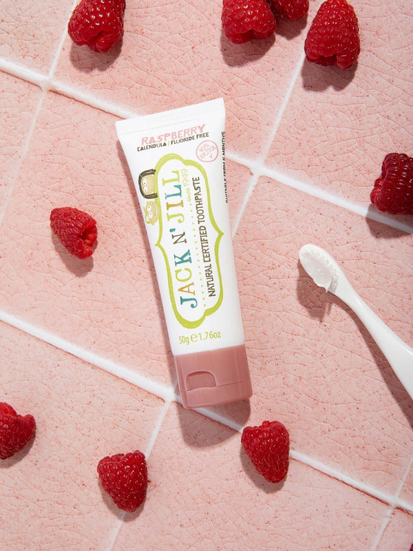 Jack N' Jill Raspberry toothpaste tube arranged on pink tiles with fresh raspberries and a white toothbrush.