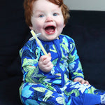 Smiling red-haired child in blue dinosaur-print pyjamas sitting on a dark sofa and holding a yellow Jack N' Jill toothbrush with a clear safety shield beside their open mouth.