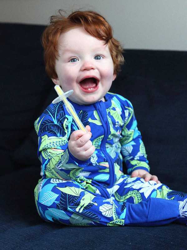Smiling red-haired child in blue dinosaur-print pyjamas sitting on a dark sofa and holding a yellow Jack N' Jill toothbrush with a clear safety shield beside their open mouth.