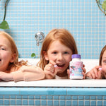 Children smiling over a blue-tiled bathtub while displaying colourful Jack N' Jill Tooth Sparkles bottles.
