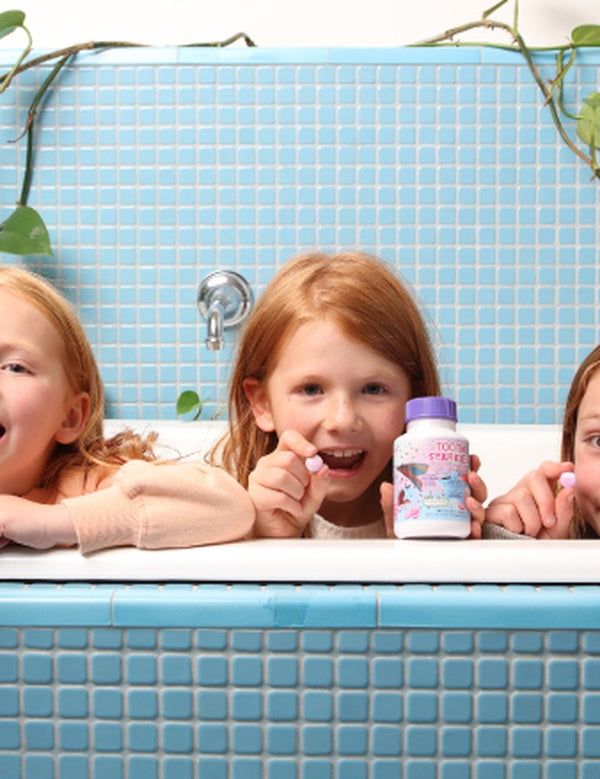 Children smiling over a blue-tiled bathtub while displaying colourful Jack N' Jill Tooth Sparkles bottles.