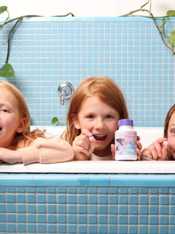 Children smiling over a blue-tiled bathtub while displaying colourful Jack N' Jill Tooth Sparkles bottles.