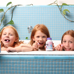 Three children leaning over the rim of a blue-tiled bathtub, smiling with Jack N' Jill Tooth Sparkles bottles lined up in front of them and green trailing plants behind.
