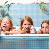 Three children leaning over the rim of a blue-tiled bathtub, smiling with Jack N' Jill Tooth Sparkles bottles lined up in front of them and green trailing plants behind.